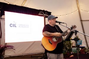 Musician performs with acoustic guitar under white tent with CIL banner displayed behind.