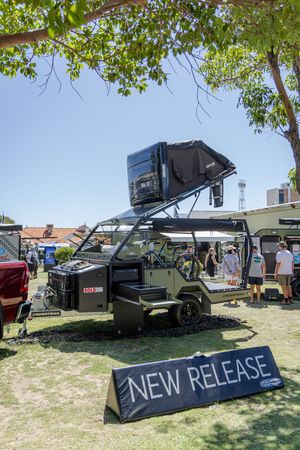A military-style off-road trailer with rooftop tent displayed at outdoor event with 'NEW RELEASE' sign beneath it.