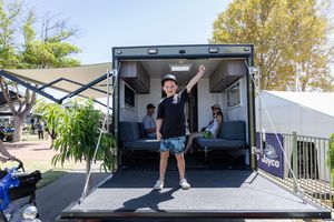 A travel trailer has its rear ramp door open with two people standing and sitting inside the cargo area.