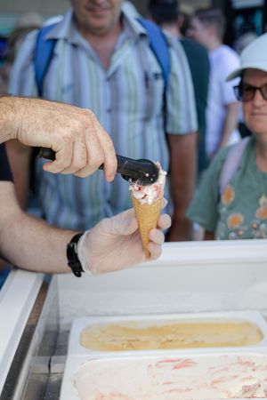 Person scooping ice cream into a cone at a vendor stand with people waiting in line.