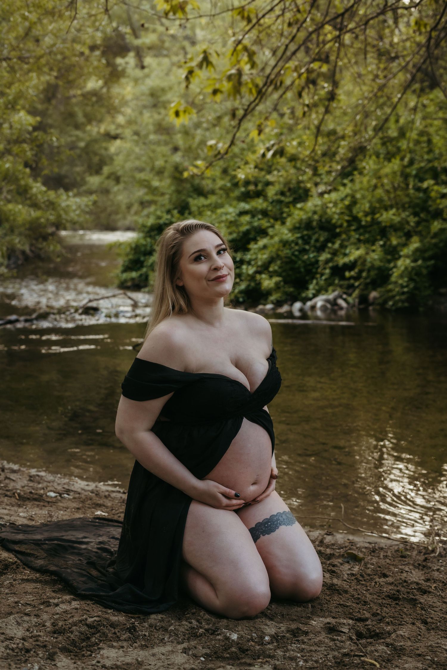 An expectant mother wearing a black off-shoulder dress stands in shallow river water surrounded by green foliage.