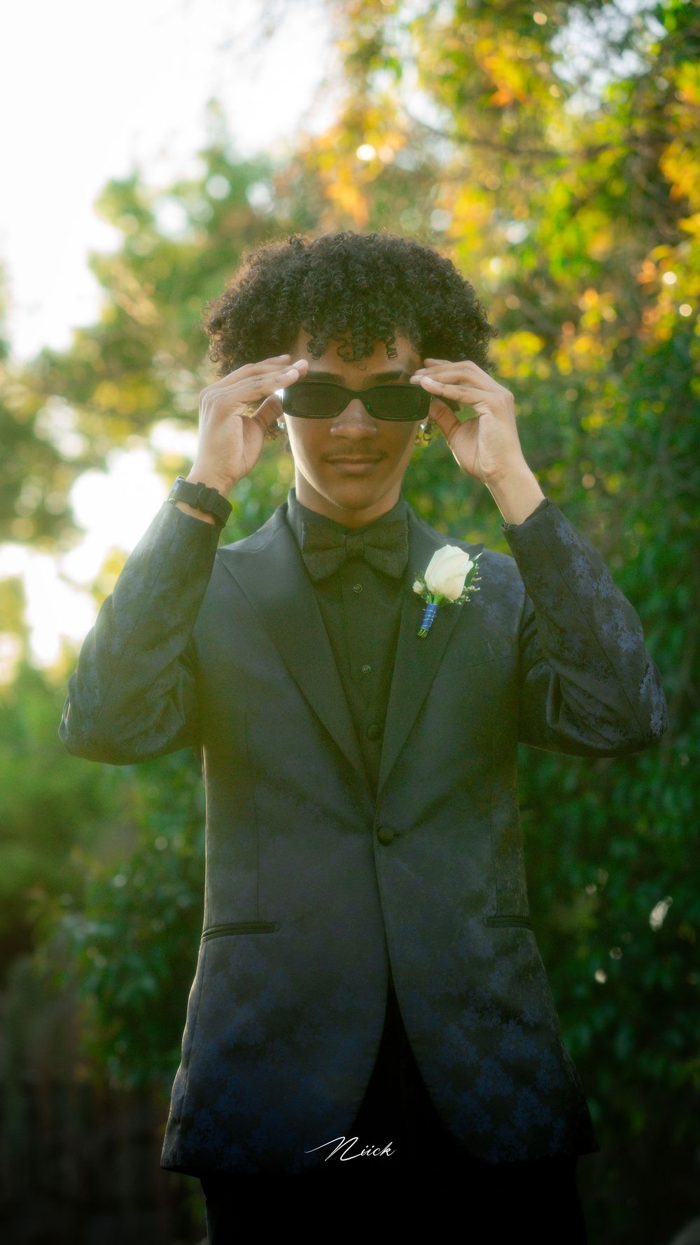 Formal portrait of someone in a black tuxedo adjusting sunglasses with a boutonniere against blurred green foliage.