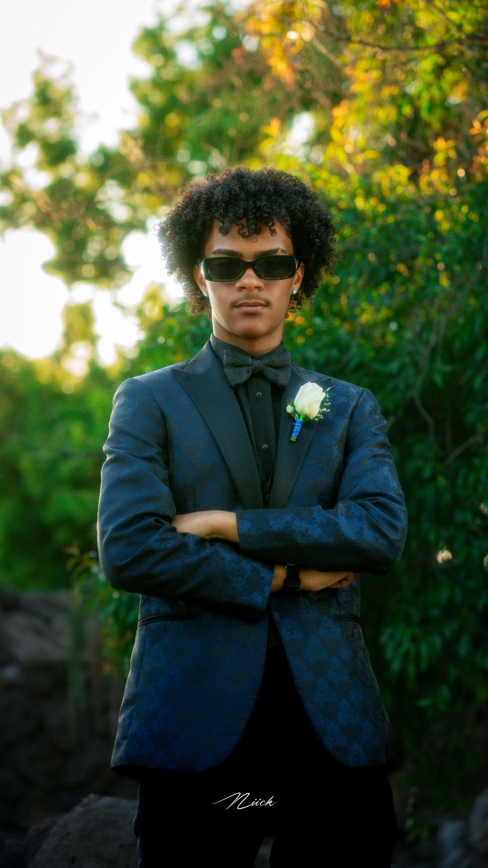 Someone in a navy blue suit with sunglasses and a white boutonniere poses with crossed arms against autumn foliage.