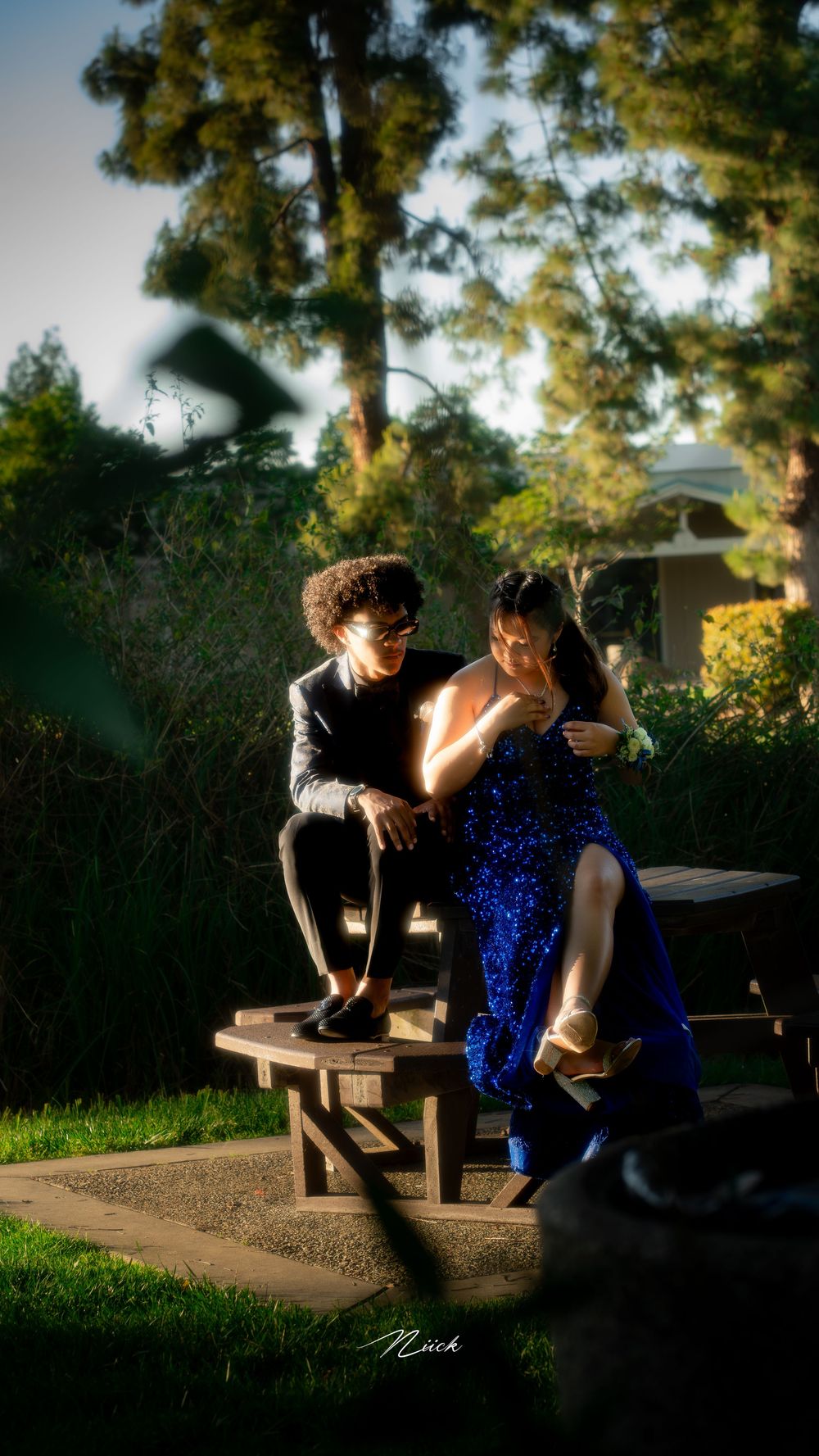 A couple sits together on a wooden bench at sunset, with one person wearing a sparkling blue dress and the other in black.