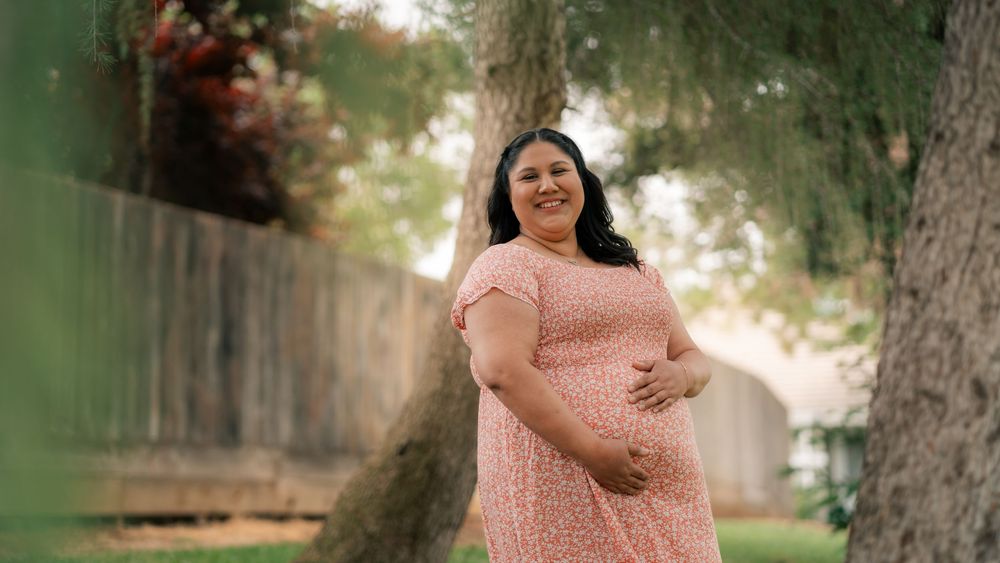 A pregnant woman in a flowing pink dress poses outdoors in a serene garden setting with sunlit trees in the background.