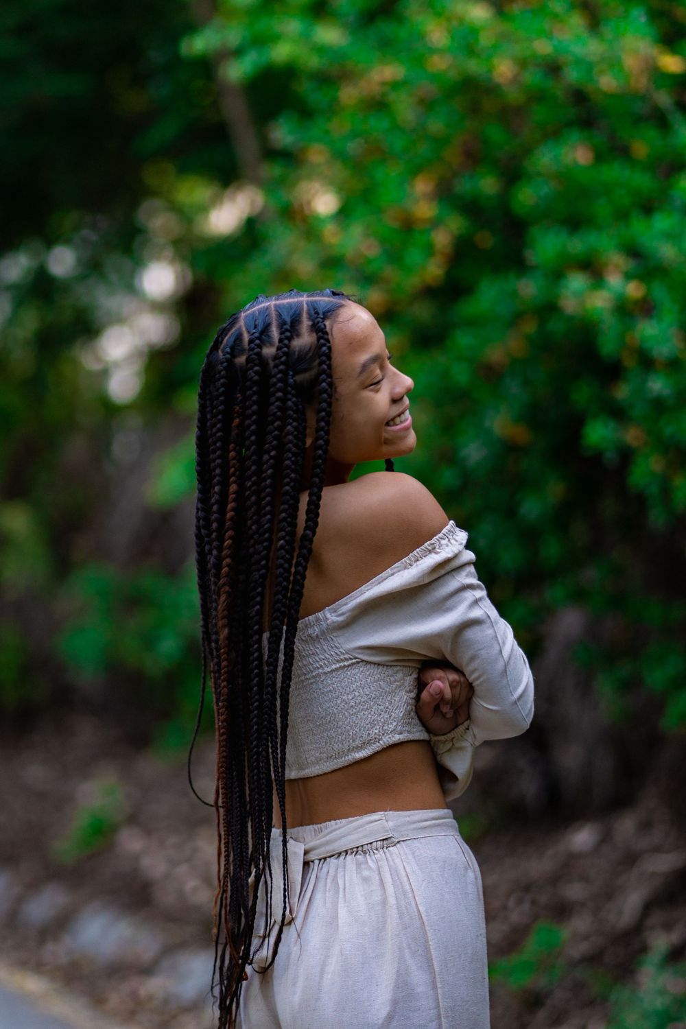 A person with long braided hair poses outdoors in a white off-shoulder top and skirt against a green natural background.