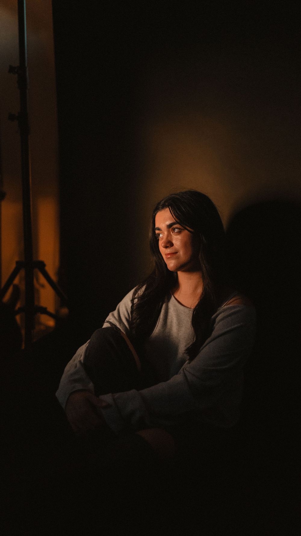 Atmospheric portrait in dark room illuminated by warm ambient lighting showing person in casual attire looking to the side.