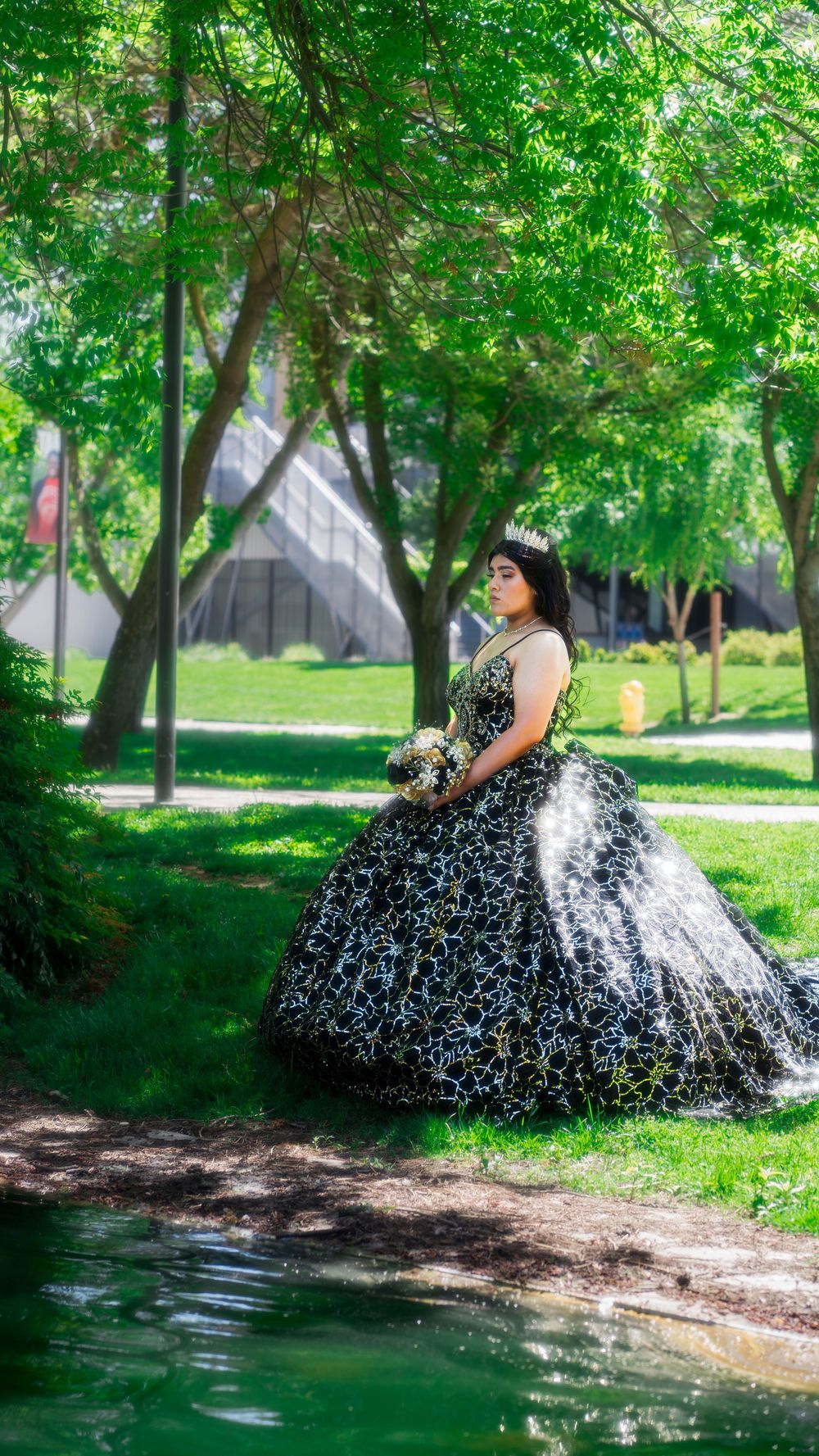 A person in an elegant black and white ball gown poses in a lush green park setting near a stream.