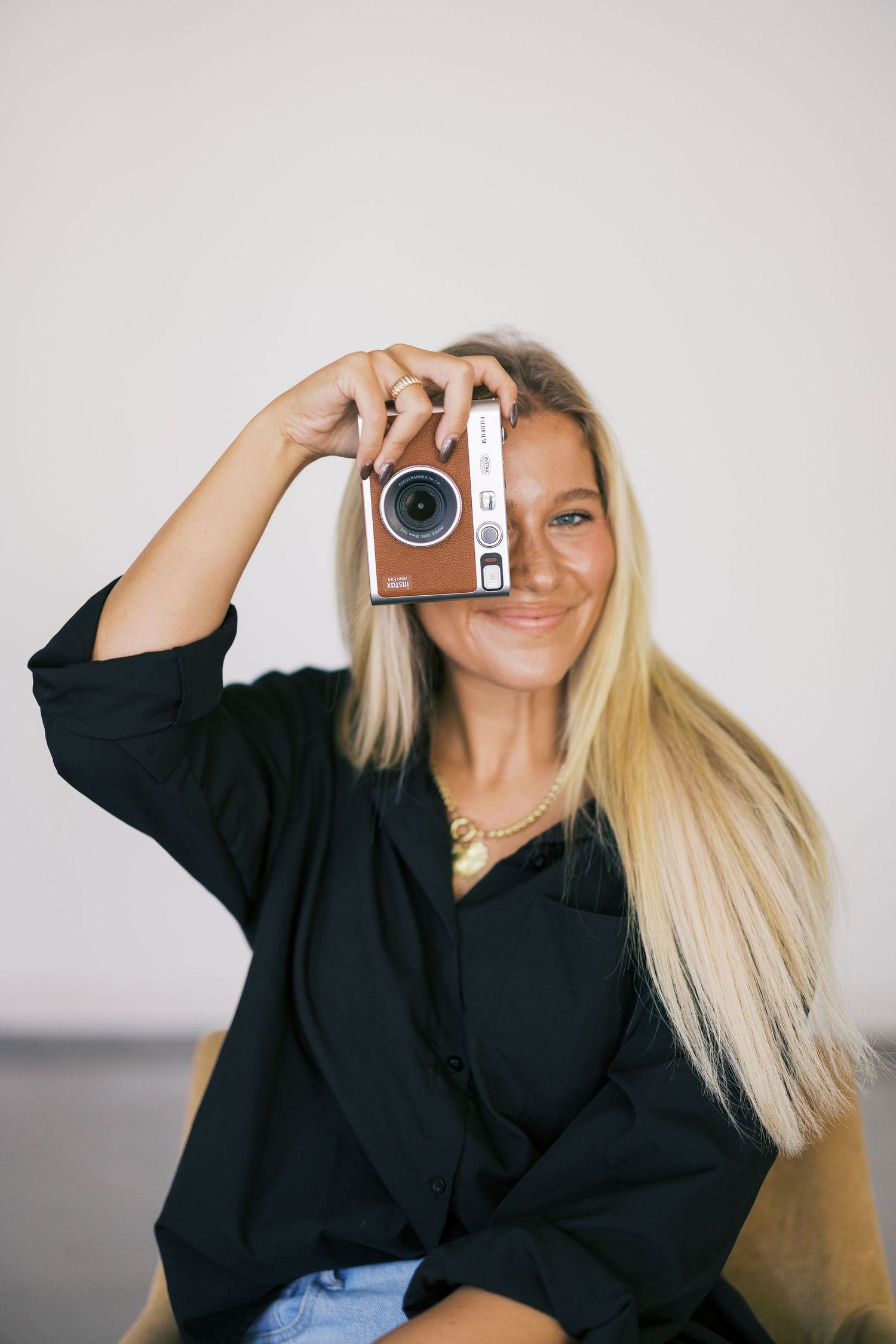 Photographer holds vintage silver camera while wearing black button-down shirt and light denim against neutral background.