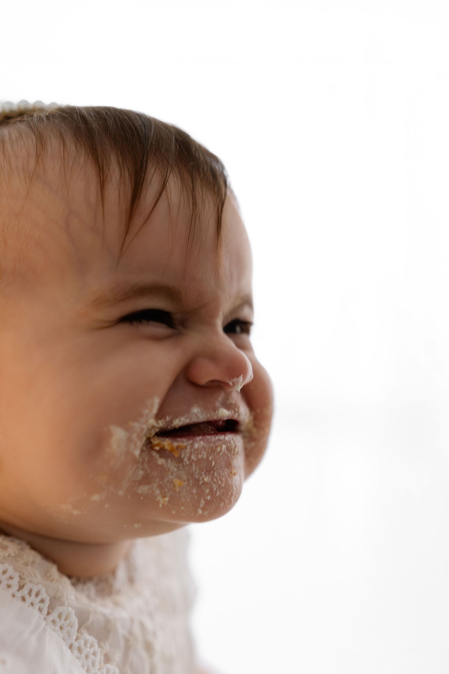 Adorable baby with messy face smiling and wearing white lace clothing against light background.