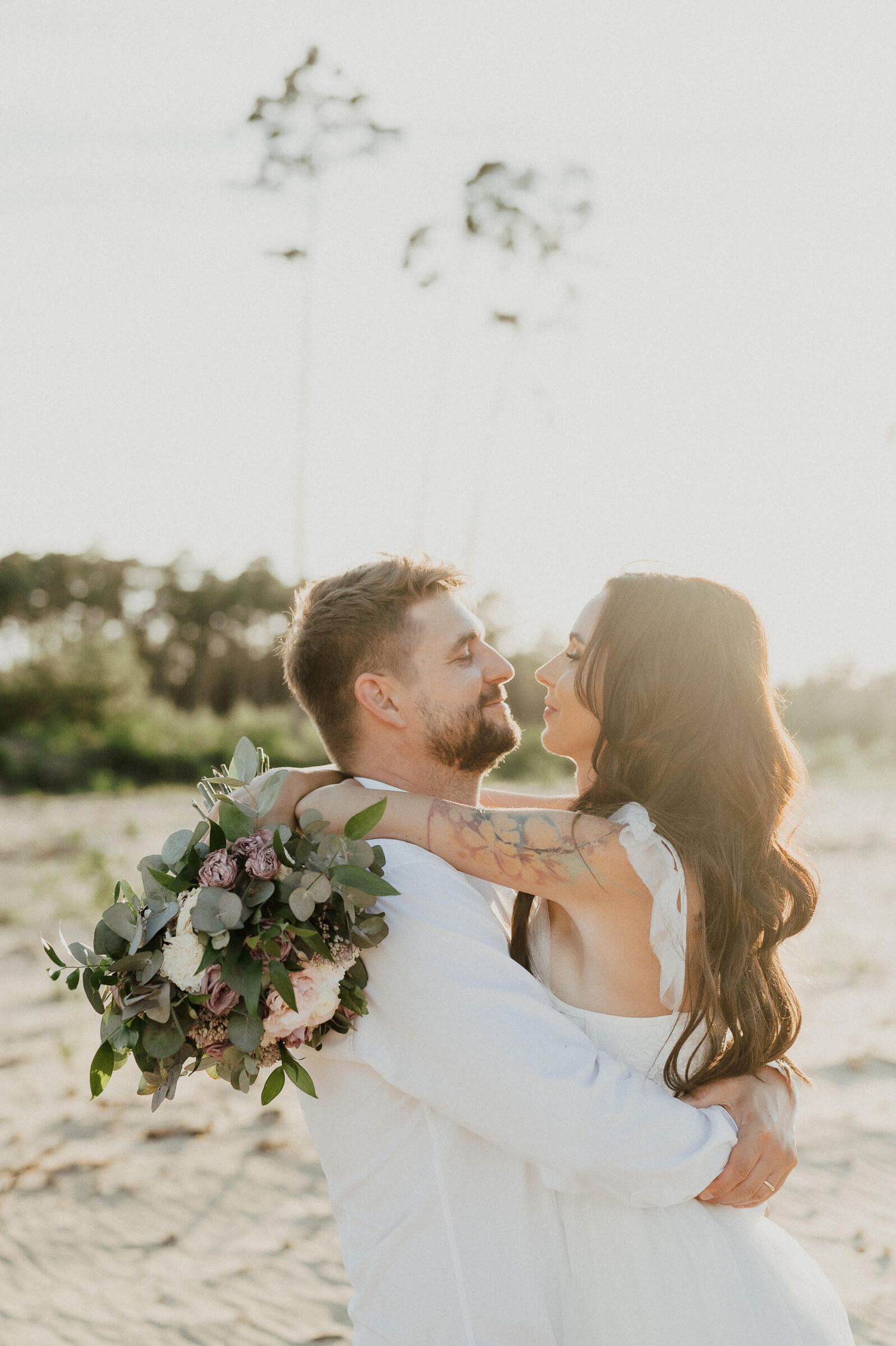Romantic beach portrait of couple embracing with floral bouquet at sunset.