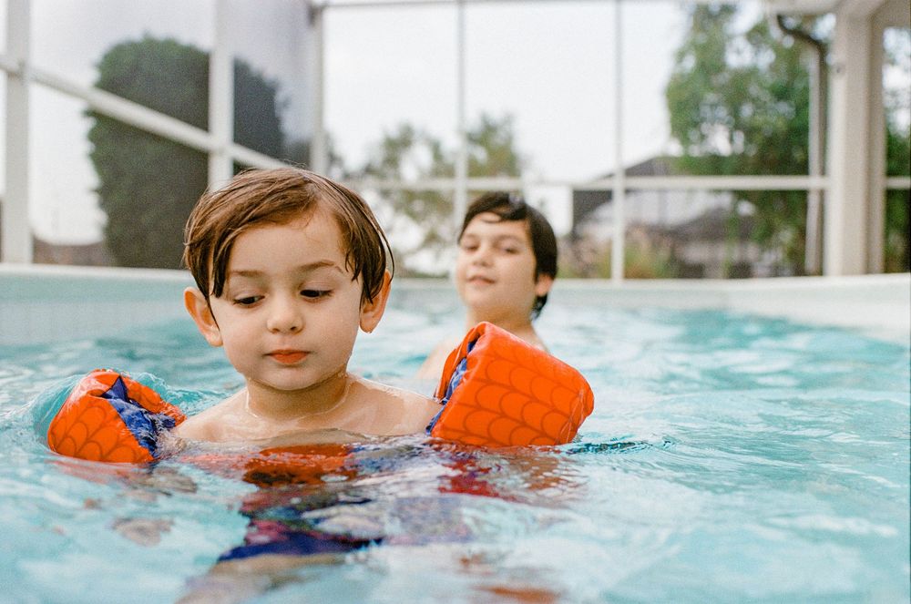 Two children swimming with orange arm floaties in a bright pool on a sunny day.