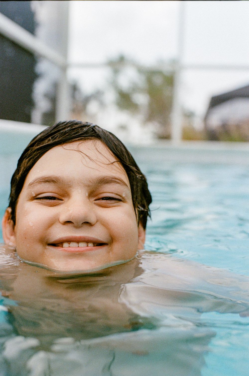 A swimmer's smiling face emerges from the rippling blue water of a swimming pool on a sunny day.
