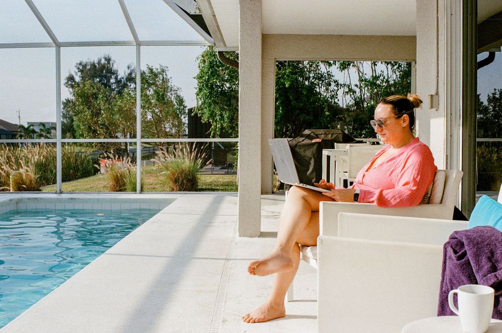 Person in coral colored shirt relaxing by poolside with laptop on a sunny day at covered patio area.