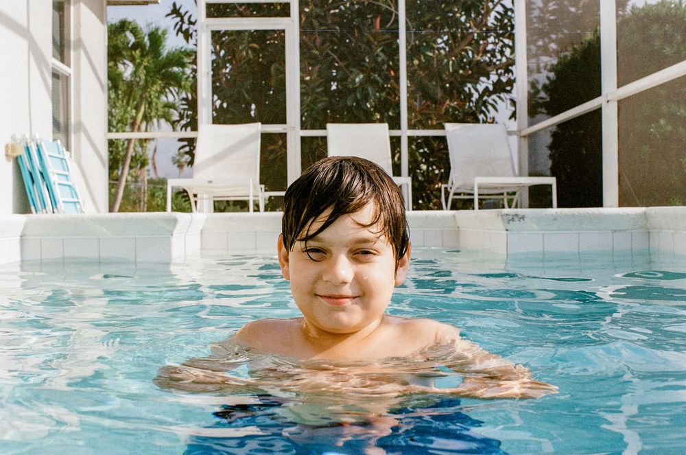 Someone swimming in a bright blue pool on a sunny day with glass windows and white furniture visible in background.