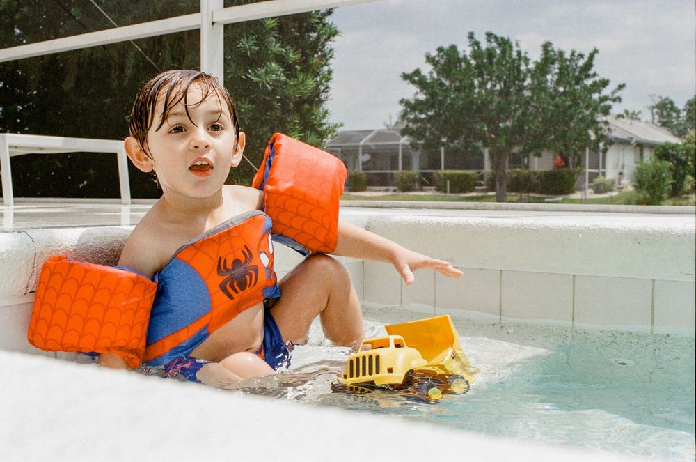 Child wearing orange water wings plays with yellow toy truck in a swimming pool on a sunny day.