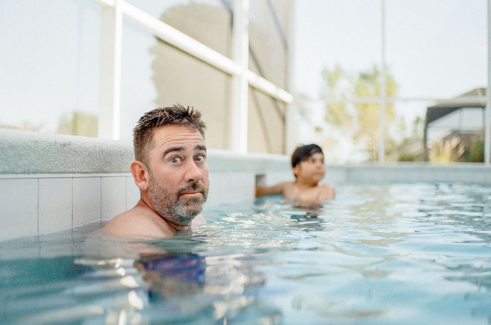 Two people relax in a bright blue swimming pool with a white architectural structure visible in the background.
