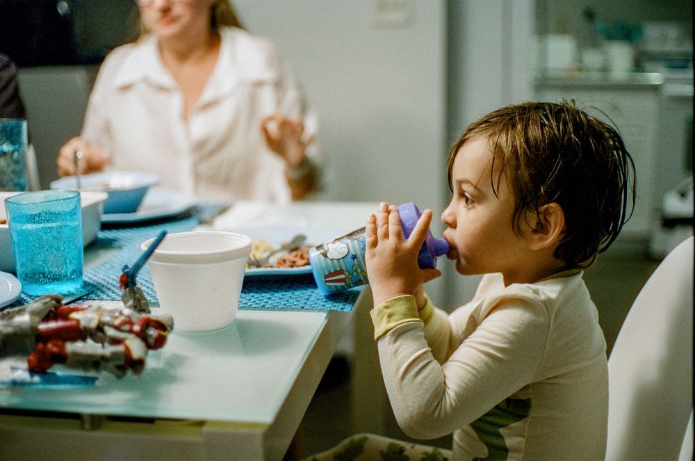 A young child drinks from a cup at a dining table with blue glassware and plates during mealtime.
