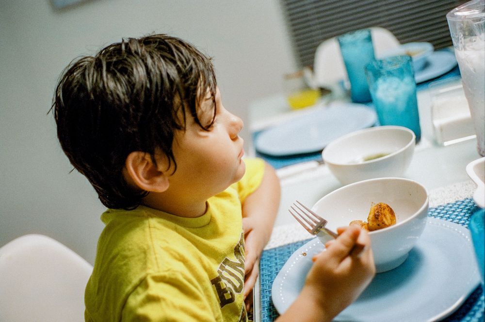 A child wearing a bright yellow shirt sits at a table with white plates and utensils during mealtime.