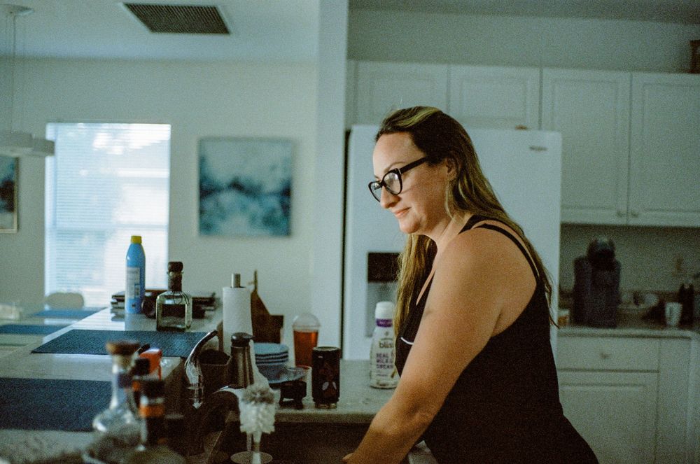 Person wearing glasses stands in a bright kitchen with glasses and bottles on the counter.