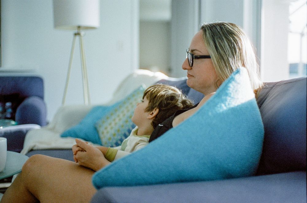 Someone relaxes on a blue couch with a small dog while using a mobile device in a sunlit living room.