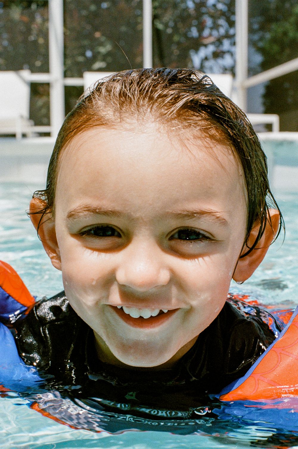 A child wearing a swim shirt and orange floaties enjoys swimming in a pool on a sunny day.