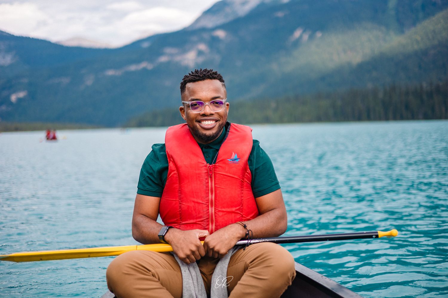 Person wearing a red life vest smiles while sitting in a canoe on a scenic mountain lake.