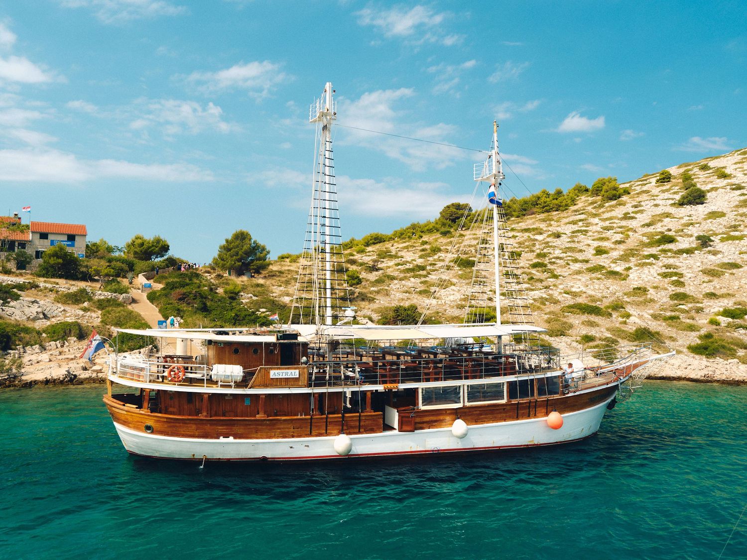 Traditional wooden tourist boat with two masts cruising in turquoise Mediterranean waters near rocky coastline.