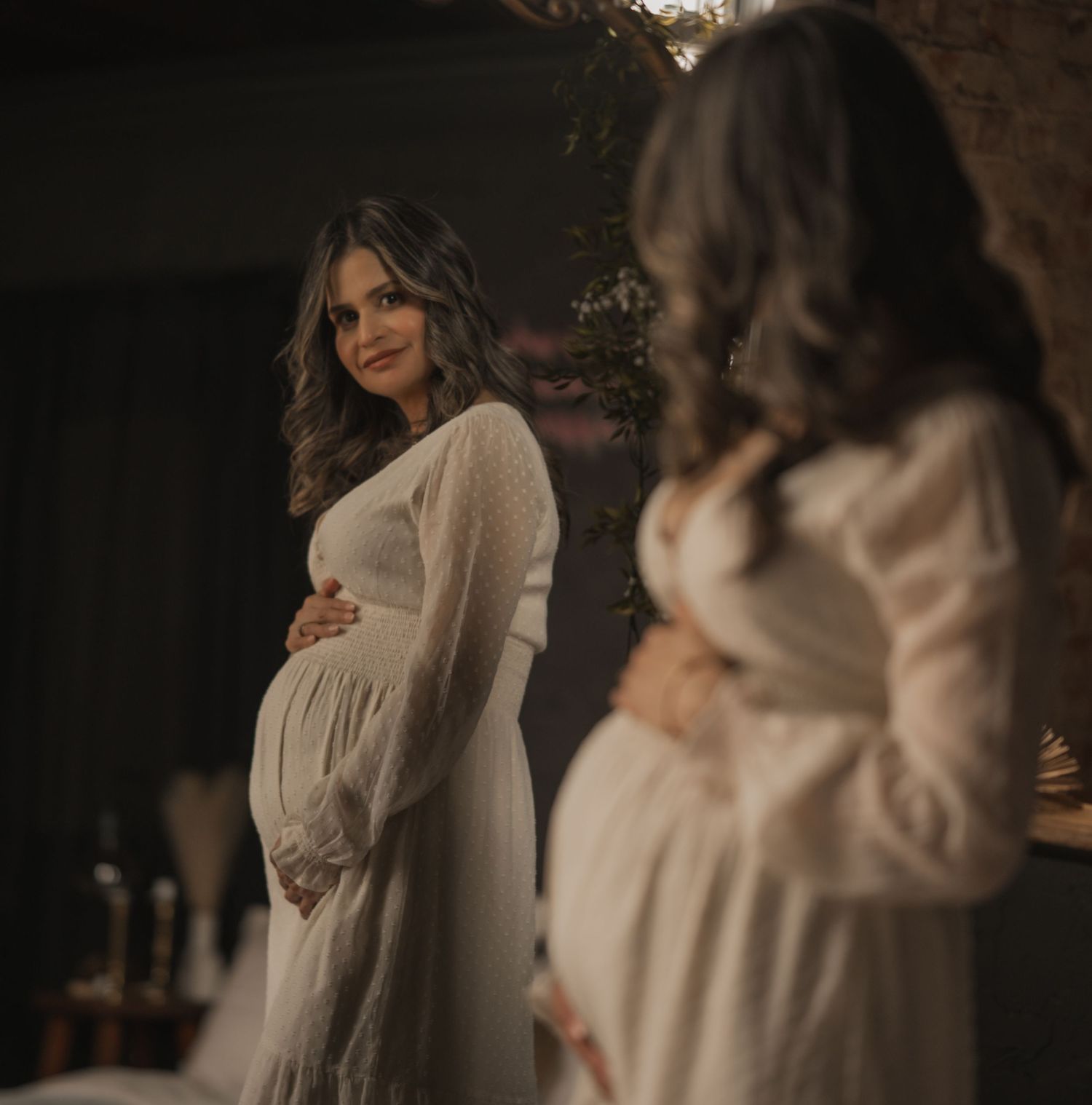 A pregnant figure in a white flowy dress stands beside a mirror in a dimly lit room creating an ethereal maternity scene.