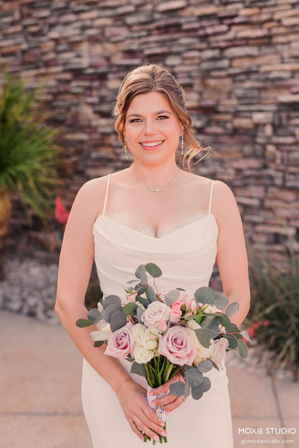 Bride holds a beautiful bouquet of pink and white roses with eucalyptus against a stone wall.