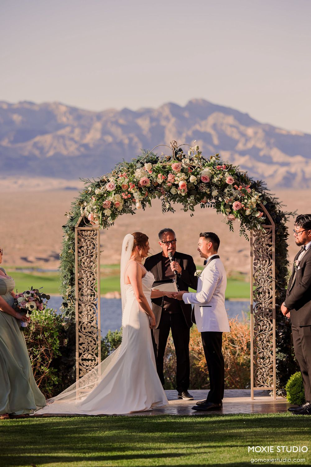 Wedding ceremony takes place under a floral arch with mountains and desert landscape in the background at sunset.