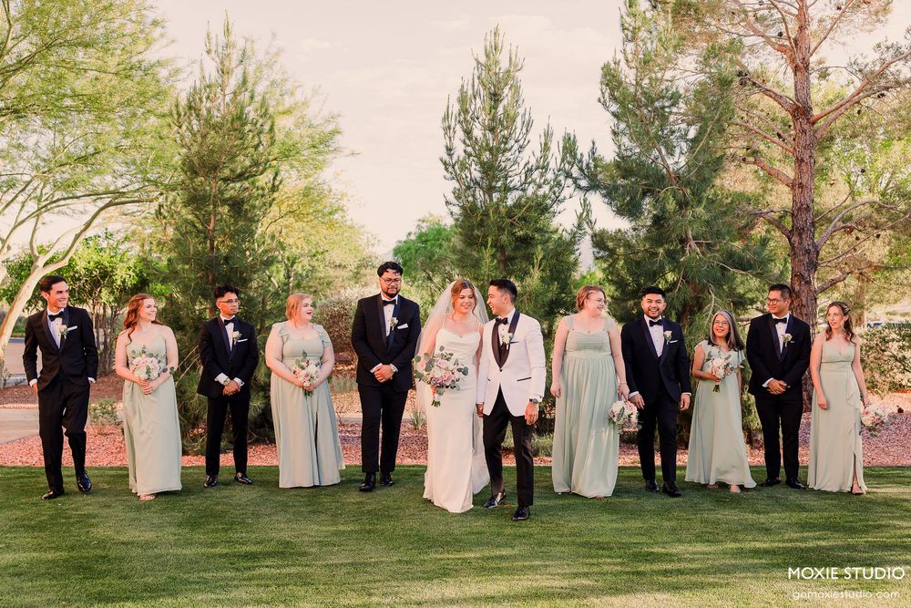 Wedding party members in pastel attire stand together on a grassy lawn during golden hour.