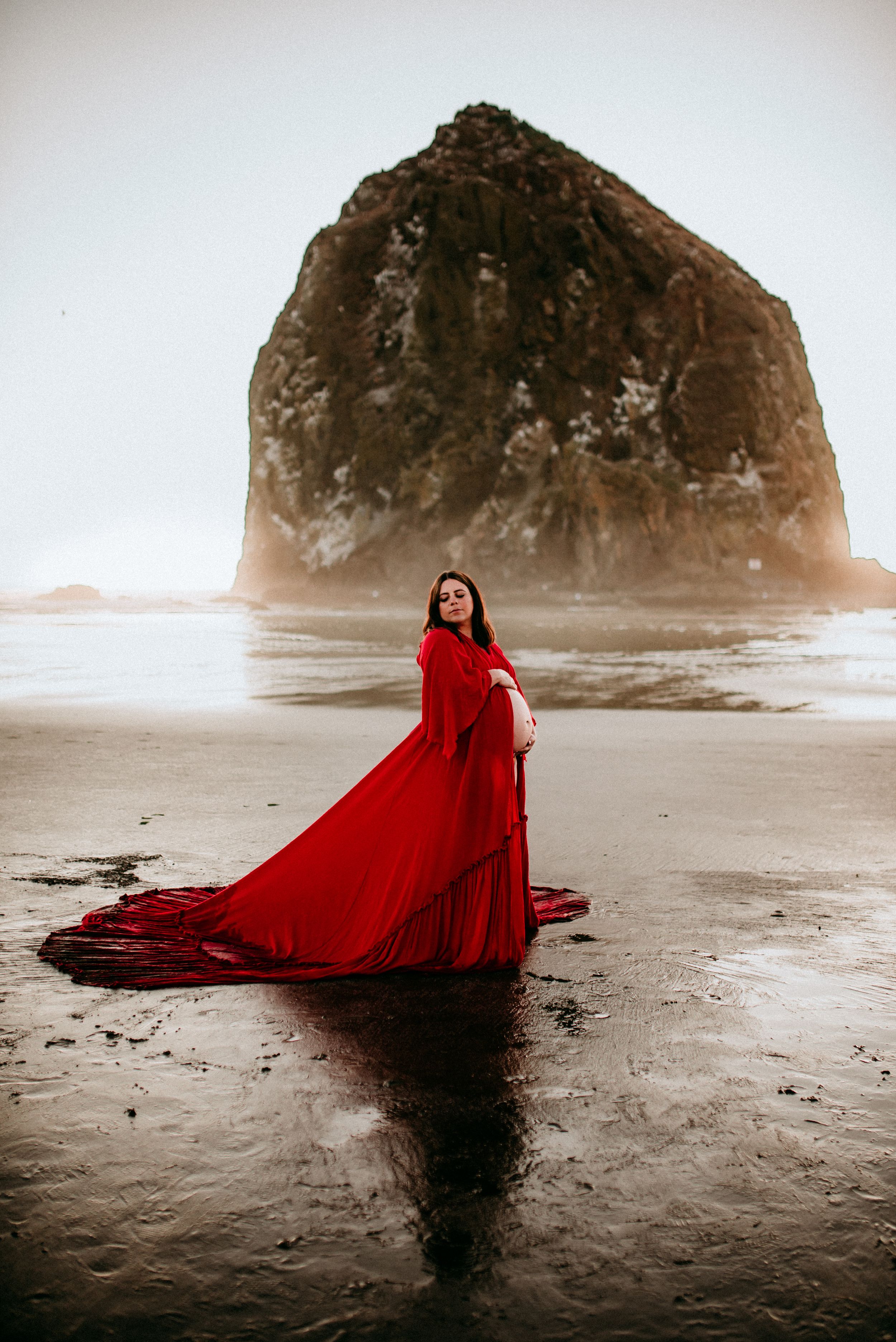 Pregnant woman with a long, flowy, red dress with her belly exposed at Canon Beach in Oregon.