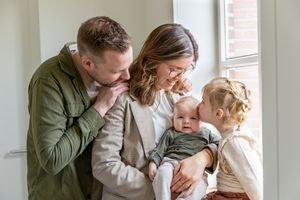 Gezin van vier met pasgeboren baby, staand bij een raam. Grote zus zit in de vensterbank en geeft haar broertje een kus, ouders kijken