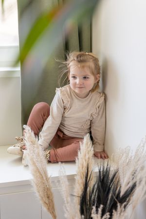 jong meisje zitten in de vensterbank, gefotografeerd vanachter een plant tijdens de newbornsessie van haar baby broertje in Haarle