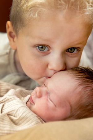 Blonde peuter geeft zijn kleine pasgeboren broertje een kus op het hoofd tijdens een babyshoot bij hen thuis in Lemelerveld