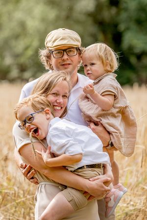 Jong gezin tijdens een gezinsfotoshoot staand in een korenveld. Ouders tillen de kinderen, ze knuffelen elkaar gehuld in wit / bruine kleren