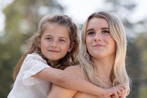Twee zusjes bij elkaar op de rug. De een blond, de ander bruin haar. Kijkend langs de camera van Fotograaf Inge Muller uit Lemelerveld