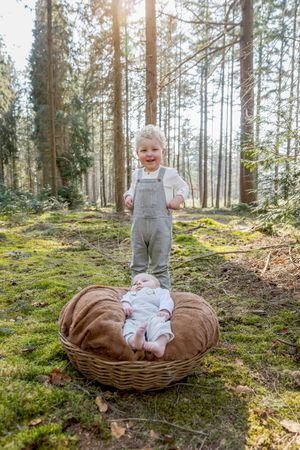 Jongetje met blonde krullen in een tuinbroek staat op de Lemelerberg, kijkend naar zijn pasgeboren zusje in een ronde rieten mand voor zich