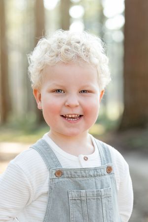 Peuter met blonde krullen. Jongetje draagt een tuin en lacht richting de camera van Fotograaf Inge Muller