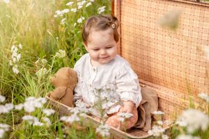 Peuter in witte blouse, zittend in een openstaande koffer met een teddybeer in haar hand in een veld met bloemen in Lemelerveld