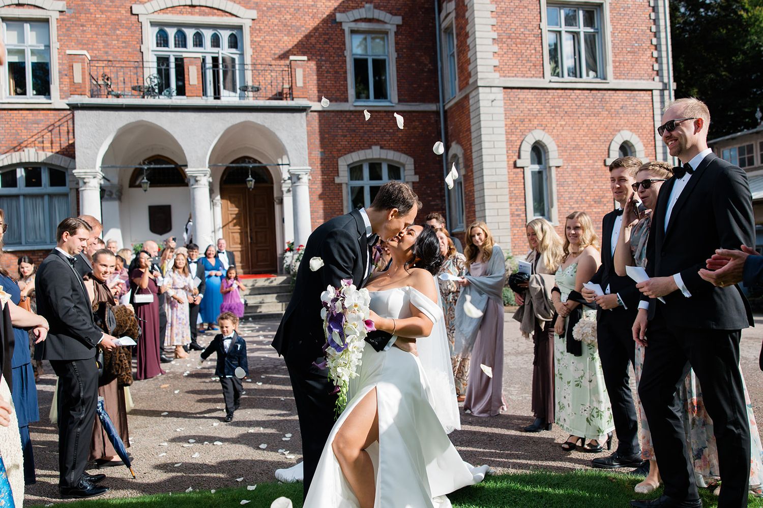 Wedding guests gather outside a brick building to celebrate as newlyweds share their first kiss as a married couple.