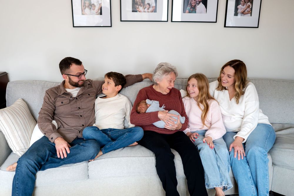 Multi-generational family relaxing on gray sofa with elderly woman holding a newborn baby.