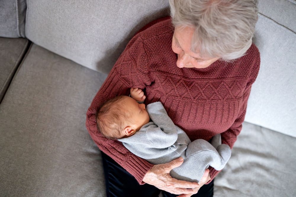 Elderly grandmother with gray hair holding sleeping newborn baby on gray sofa.