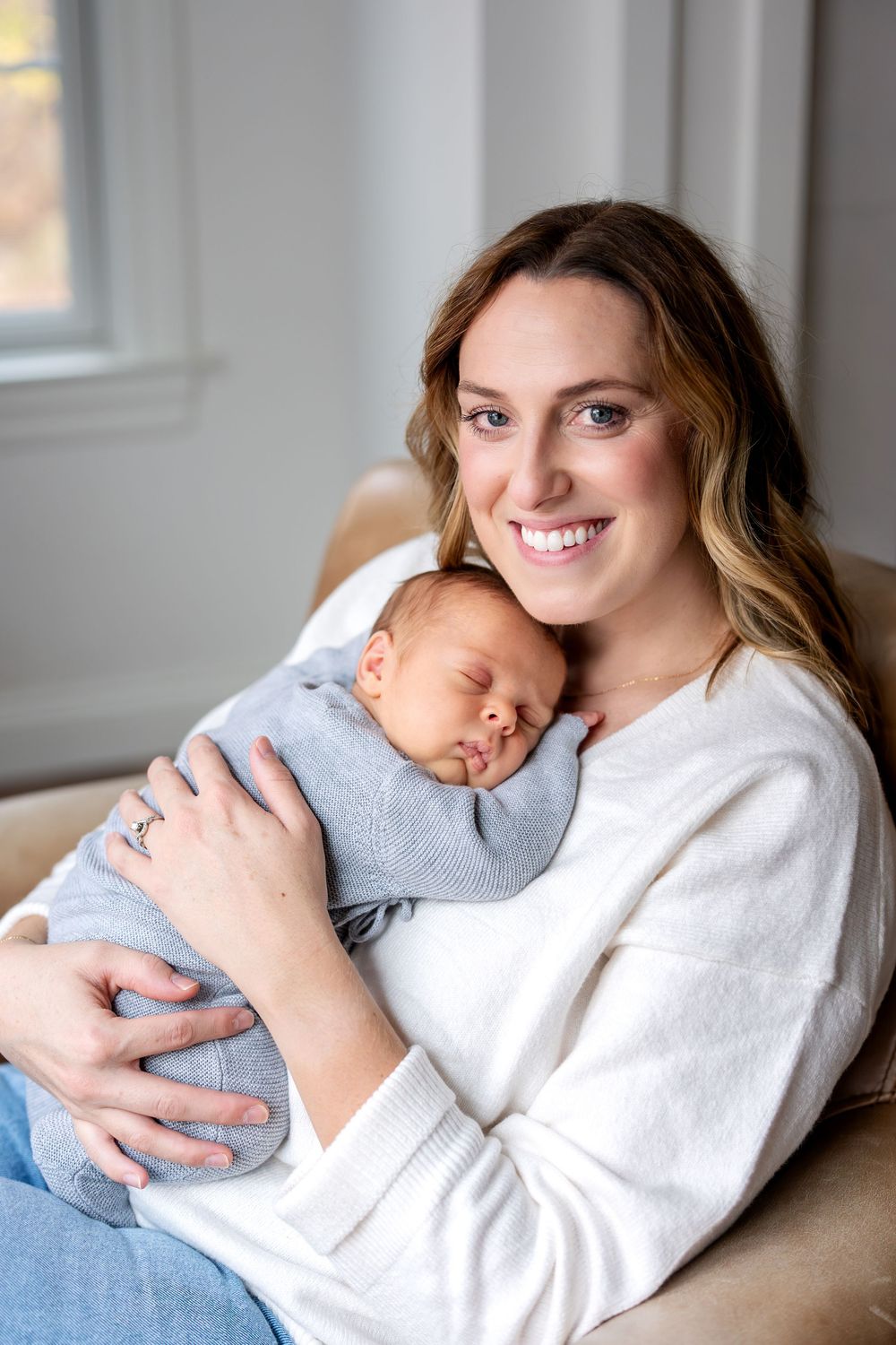 Smiling mother holding sleeping newborn baby in white sweater in chair.