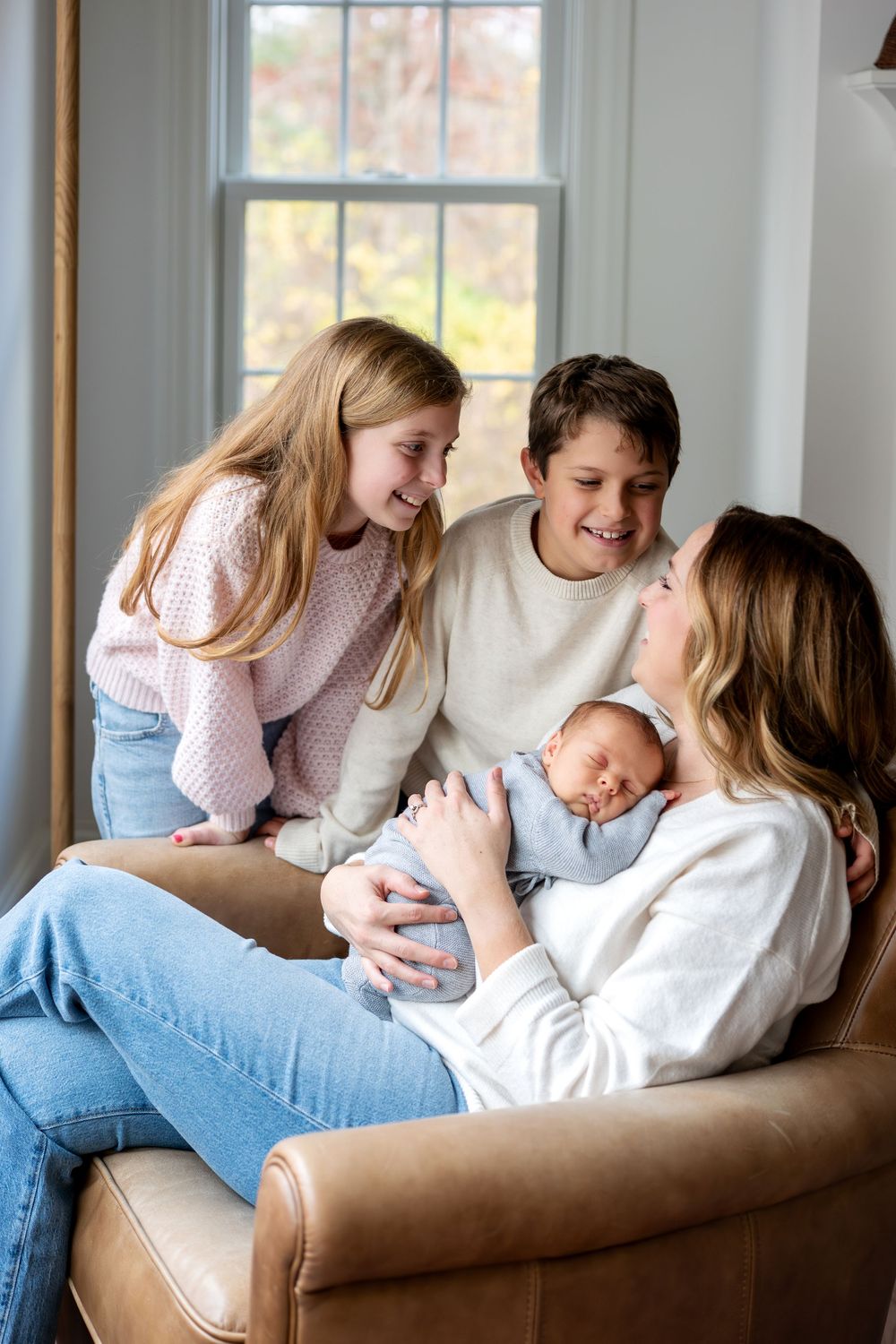 A mother sits in a leather chair holding her newborn baby while two older siblings smile adoringly at the infant.
