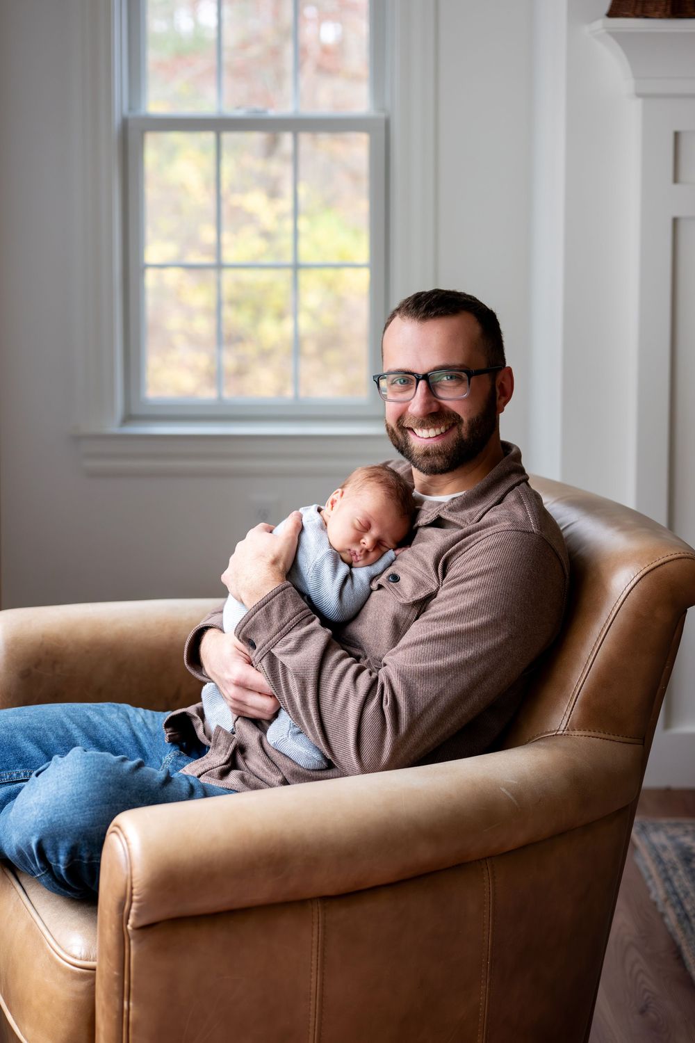 A smiling father wearing glasses holds his sleeping newborn baby while sitting in a leather armchair.