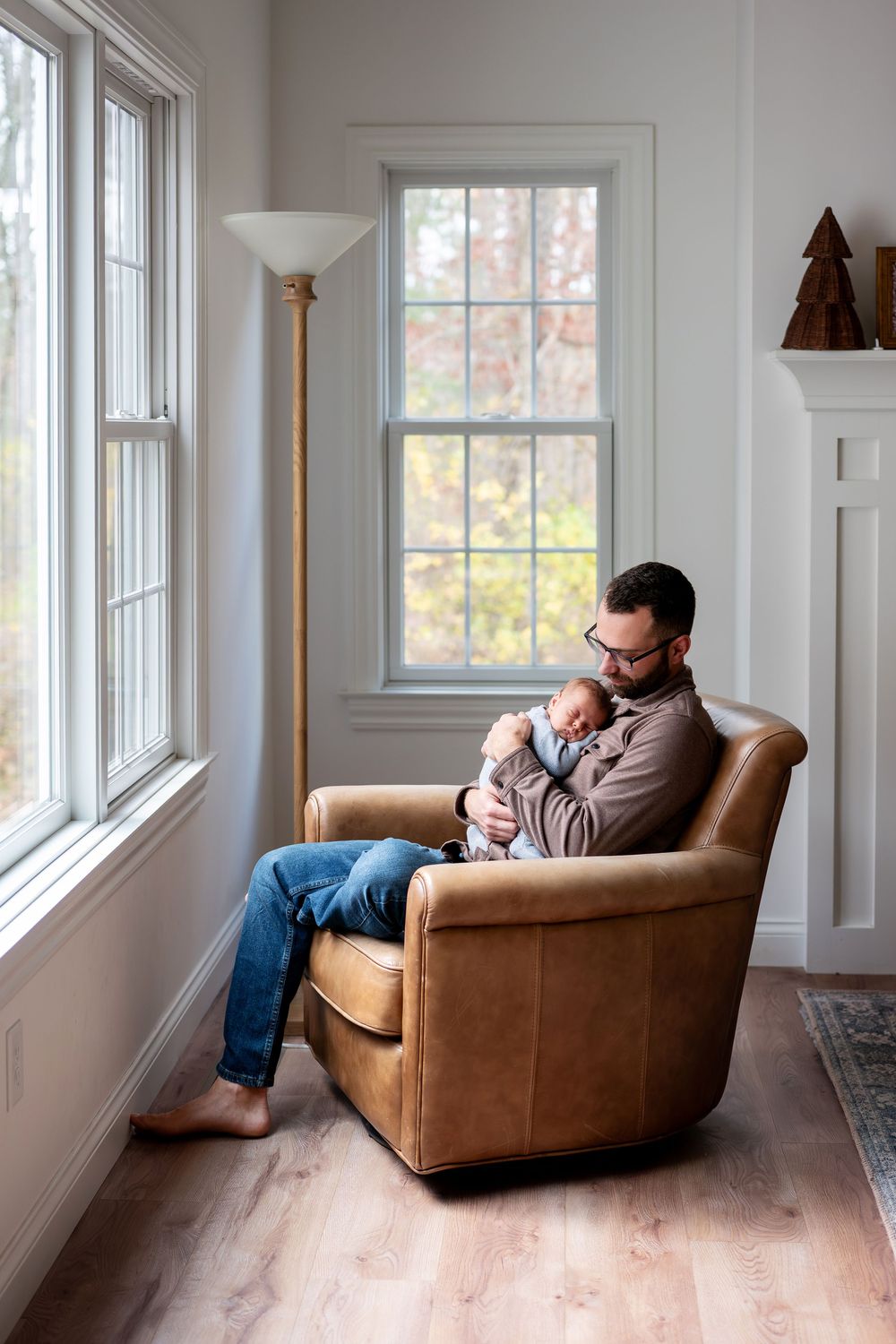 A father sits barefoot in a leather armchair by windows holding his sleeping newborn in a cozy home setting.