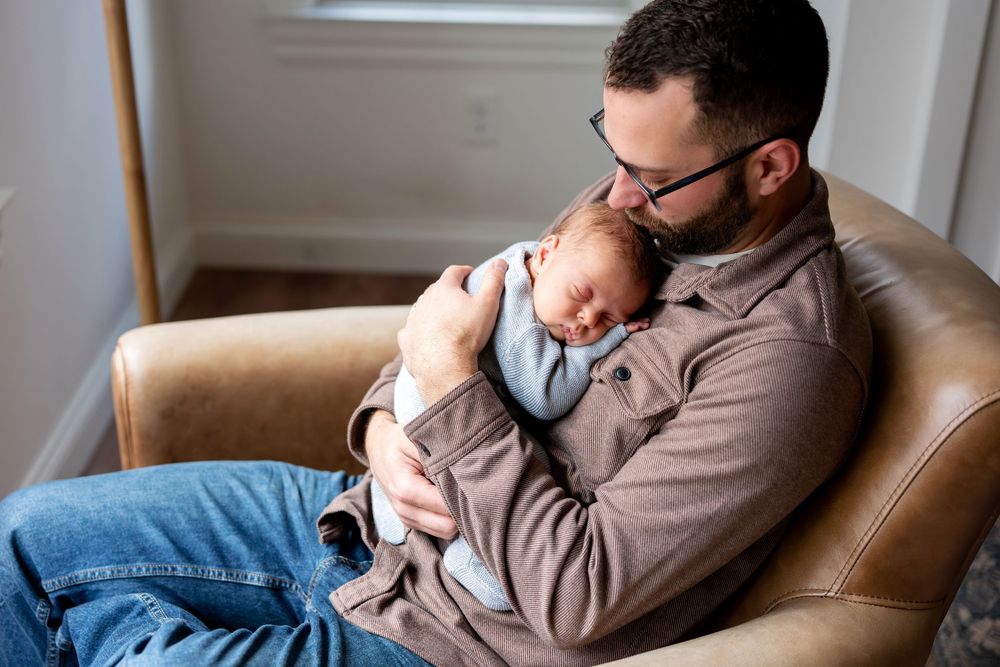A close-up of a father kissing his peacefully sleeping newborn baby dressed in blue while seated in a chair.