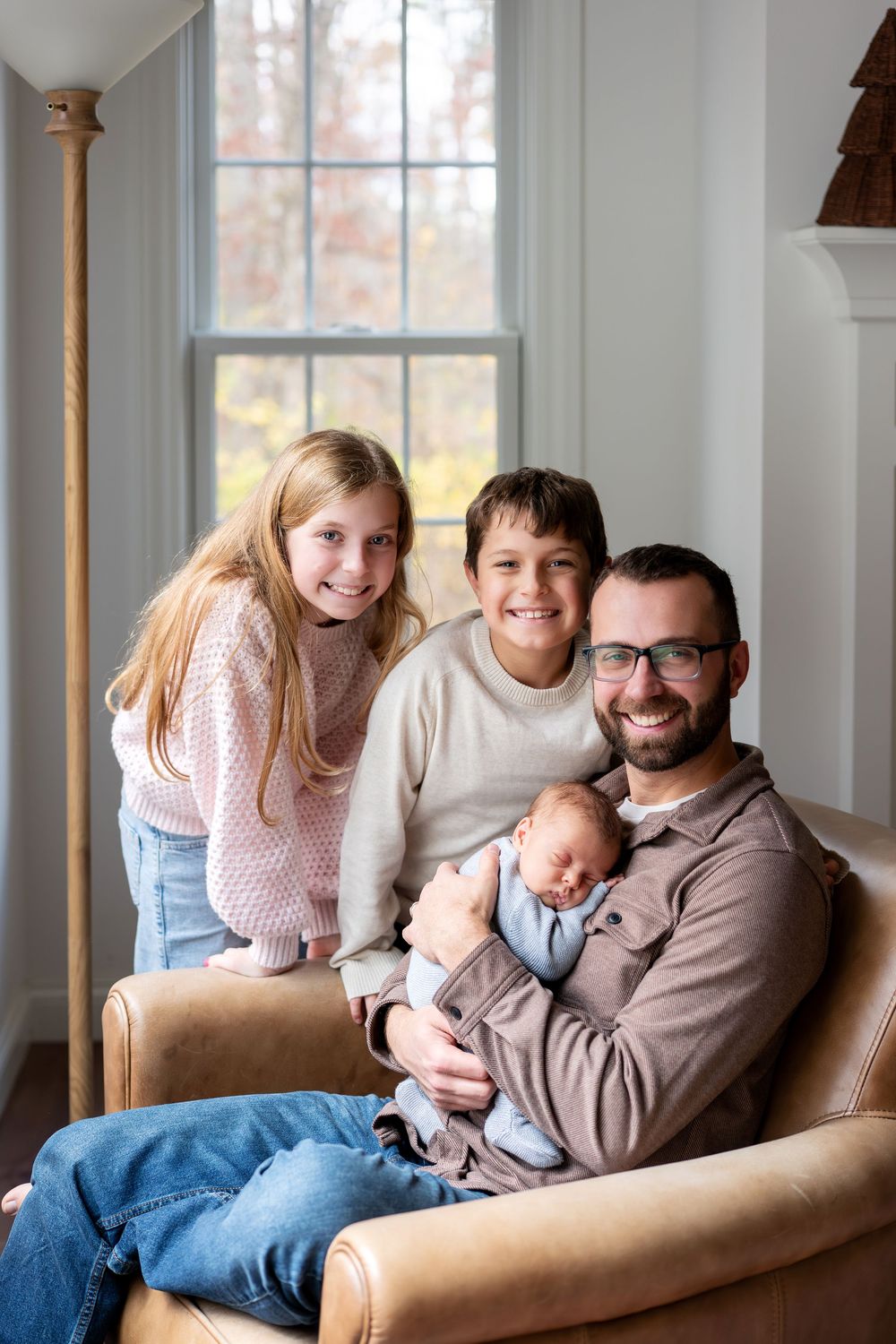 A father with glasses holds his newborn while two smiling older children pose together in a leather armchair.